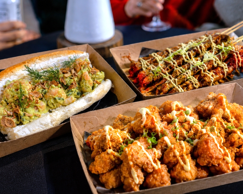 Three open containers filled with finger food including cauliflower bites, chicken skewers and a prawn and avocado bahn mi roll. The food is placed onto a table with a black table cloth