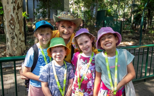 Four kids wearing colourful summer clothes, a bucket hat and lanyard with an elderly woman in a yellow top and a decorated stetson hat all smiling to the camera. Behind them is a dark green rail, stone path and green trees