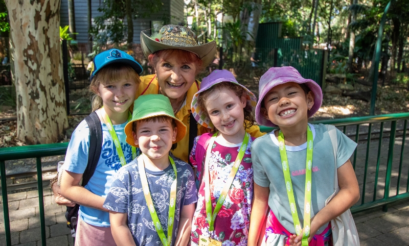Four kids wearing colourful summer clothes, a bucket hat and lanyard with an elderly woman in a yellow top and a decorated stetson hat all smiling to the camera. Behind them is a dark green rail, stone path and green trees