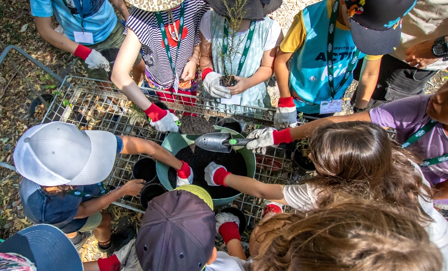 A birds eye view of a group of kids standing around a pot filled with soil, wearing garden gloves. They are taking soil from the main pot to put into a smaller one, some using their hand some using a small shovel.