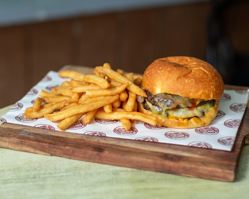 A beef burger on a panini with melted cheese next to a pile of thick cut fries served on a wooden board