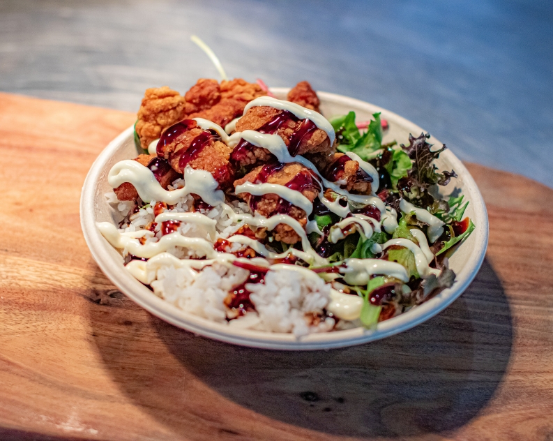 A Karaage chicken rice bowl with fresh mixed lettuce, topped with mayo and sweet and sour sauce, served on a wooden board