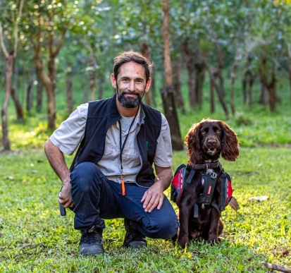 A man crouching down besides a chocolate brown dog sitting. They are both looking at the camera and are on a green grass field with trees behind them