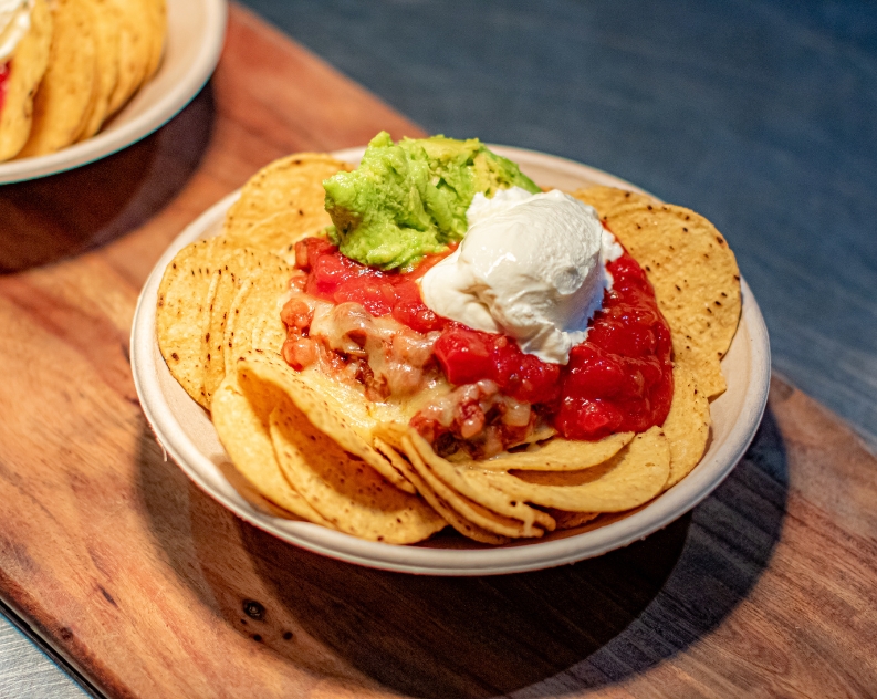 A bowl of nacho chips topped with beans, melted cheese, salsa, guacamole and sour cream served on a wooden board