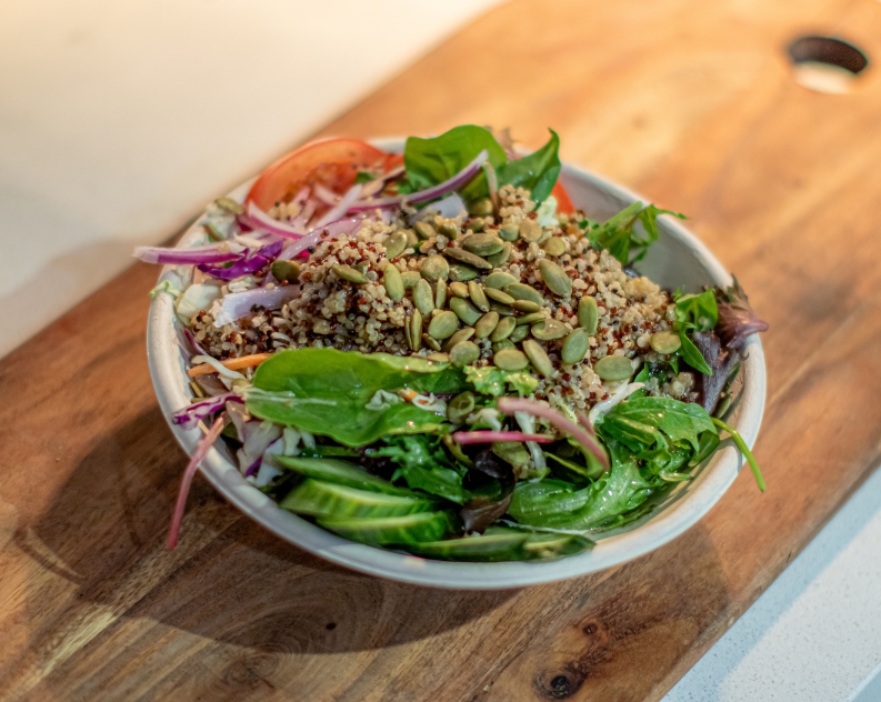 A quinoa salad with fresh lettuce, cucumber, tomato and red onion slices, topped with pumpkin seeds and served on a wooden board