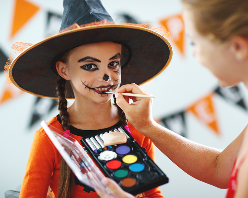 Girl in a witch hat having skeleton face paint brushed on, holding a palette of face paints, with orange and black Halloween bunting behind her